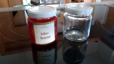Ein Glas mit Erdbeermarmelade steht neben einem leeren Glas auf einer glatten, schwarzen Arbeitsfläche in einer modernen Küche.