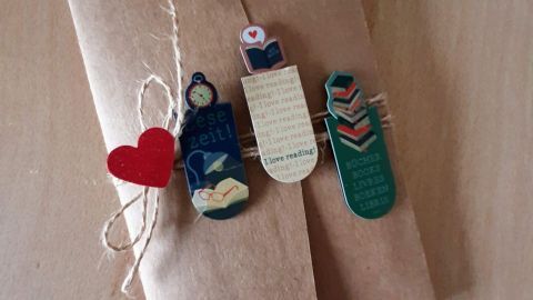 Eine dekorative Karte aus braunem Papier, liebevoll verziert mit Lesezeichen und einem roten Herz, liegt auf einem Tisch. Transkription: "Diese Zeit! I love reading! Bücher leben."