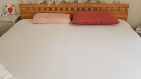 Ein großes, weißes Spannbettlaken liegt auf einem Doppelbett mit zwei Kissen, einem Nachttisch und einem Fenster im Hintergrund.