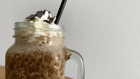 Ein Frappuccino mit Sahne und Schokoladenstückchen steht auf einem Tisch vor einer weißen Wand, bereit zum Genießen.