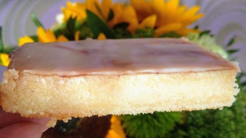 Ein saftiges Stück schwedischer Marzipankuchen wird gehalten, umgeben von bunten Blumen im Hintergrund.