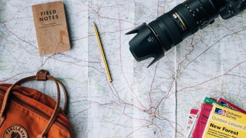 Ein orangefarbener Rucksack und eine Kamera liegen auf einer Landkarte, während ein Notizbuch und ein Bleistift bereitliegen.
