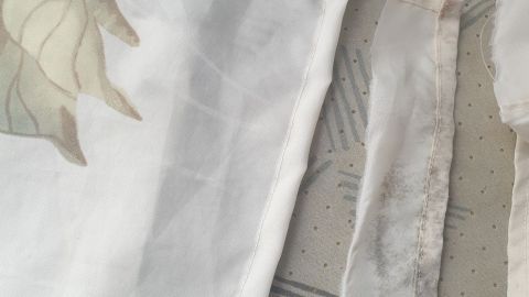 Ein Duschvorhang mit Schimmel am Saum liegt auf einer gemusterten Oberfläche, während er leicht gefaltet ist.