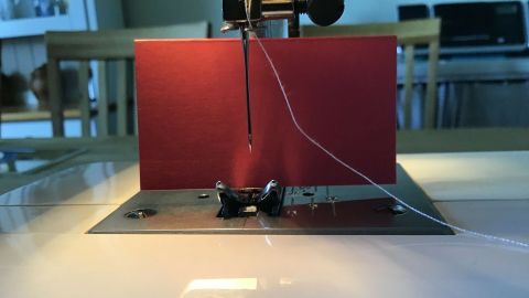 Eine Nähmaschinennadel durchfädelt ein Nadelöhr, während eine rote Karte im Hintergrund leuchtet.