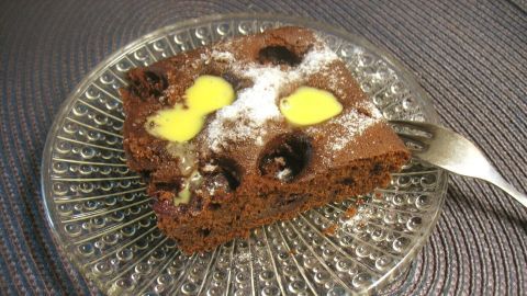 Ein Stück Kirsch-Schoko-Kuchen liegt auf einem transparenten Dessertteller, leicht glasiert und mit Butterstückchen versehen.