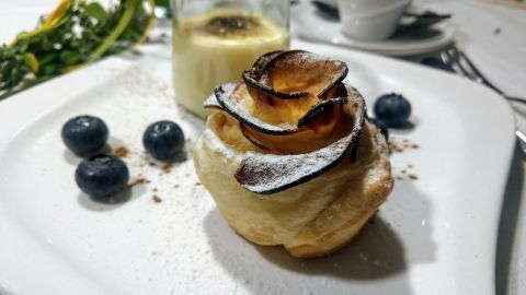 Ein zartes Dessert in Form einer Apfelrose aus Blätterteig liegt auf dem Teller, umgeben von Heidelbeeren und Sahne.