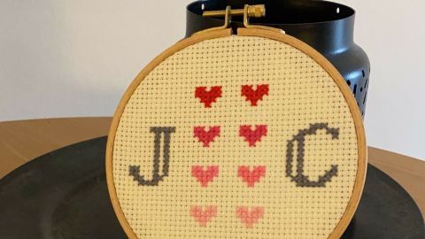 Ein gestickter Rahmen mit den Buchstaben J und C sowie Herzmotiven steht auf einem Teller, umgeben von einem Pflanzentopf.