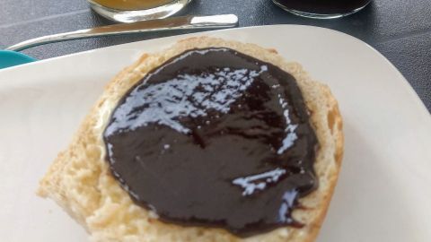 Ein Brötchen liegt auf einem Teller, belegt mit Pflaumenmus, im Hintergrund frischer Orangensaft und weitere Speisen.