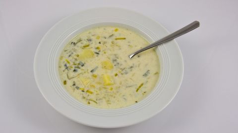 Eine cremige Lauch-Käse-Suppe mit stückigem Gemüse und einem Löffel, serviert in einer weißen Schüssel auf hellem Hintergrund.