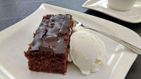 Ein Zucchini-Schokoladenkuchenstück liegt neben einer Kugel Vanilleeis auf einem weißen Teller, während eine Tasse Kaffee im Hintergrund steht.