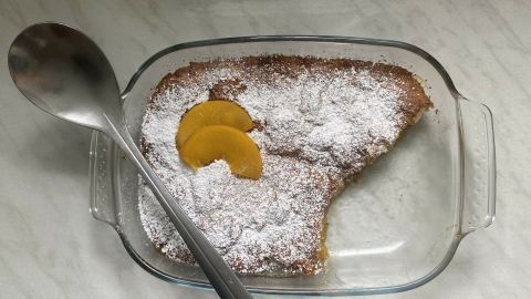 Ein Pfirsich-Cobbler mit Puderzucker und Pfirsichscheiben wird in einer Glasform auf einer Arbeitsfläche präsentiert.