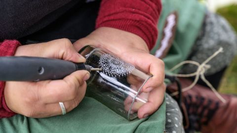 Eine Hand graviert ein Glas mit einem filigranen Muster, während die Person auf einem Stuhl im Freien sitzt.
