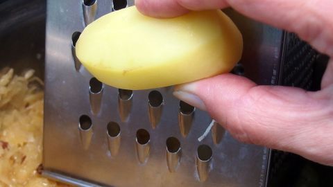 Eine Hand reibt eine Kartoffel auf einer Reibe über einer Schüssel mit Sauerkraut, um das Gericht schmackhafter zu machen.