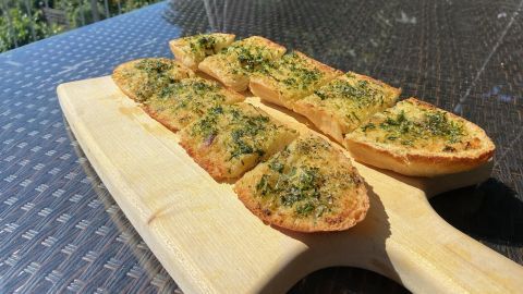 Knoblauchbrot liegt auf einem Holzbrett, frisch gebacken und mit grünen Kräutern bestreut, in einem sonnigen Garten.