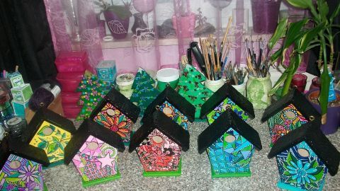 Bunte Vogelhäuser stehen auf einer Arbeitsfläche, kreativ mit Acrylfarben bemalt, umgeben von Malutensilien und Blumen.