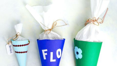 Bunte Schultüten stehen in Reihe, gefüllt mit weißem Papier, verziert mit Namen und Blumenmotiven, auf einem neutralen Hintergrund.