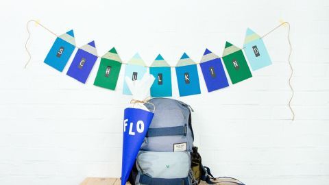 Eine bunte Girlande mit "SCHULKIND" hängt über einem Rucksack und einer Schultüte, die auf einem Tisch stehen.