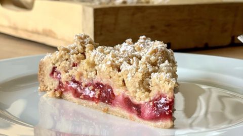 Ein Stück Streuselkuchen liegt auf einem weißen Teller, umgeben von Holz und sanften, grauen Akzenten im Hintergrund.