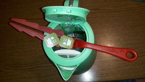 Mithilfe von einem Plastikgriff eines Staubwischers fallen Teebeutel nicht in die Kanne. Eine grüne Teekanne steht offen mit roten Zangen, die Teebeutel halten, um ein Herunterfallen beim Tee kochen zu verhindern.