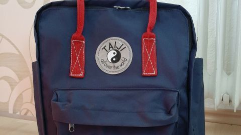 Blauer Rucksack mit roten Griffen steht auf einem Holzfußboden vor einer gemusterten Wand und strahlt Handmade-Charakter aus.