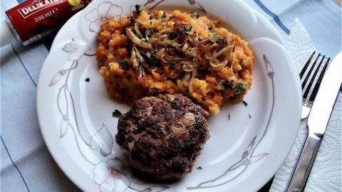 Ein Teller mit Möhren-Kartoffelstampf und einer Fleischbeilage liegt auf einem Tisch, umgeben von Besteck und Senf.