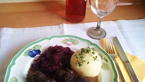 Hirschrouladen liegen auf einem Teller mit Rotkohl und Kartoffeln, während ein Roséwein im Hintergrund steht.