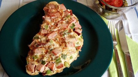 Ein Leberkäs-Omelett liegt auf einem grünen Teller, umgeben von Besteck und einem Salat aus Tomaten und Gurken.