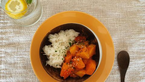 Ein Schüssel Kürbisgulasch mit Reis und Kräutern steht auf einem gedeckten Tisch, daneben ein Glas Wasser mit Zitrone.
