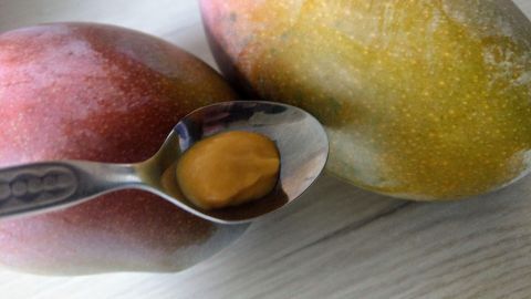 Zwei reife Mangos liegen auf einem hellen Tisch, während ein Löffel mit einer Portion Mangosenf daneben liegt.