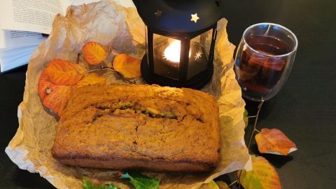 Saftiger Kürbiskuchen liegt auf Papier, umgeben von autumnalen Blättern, einer Laterne und einer Tasse heißem Getränk.