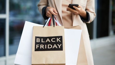 Eine Person hält Einkaufstüten mit der Aufschrift „BLACK FRIDAY“ und schaut auf ihr Smartphone vor einem modernen Geschäft.