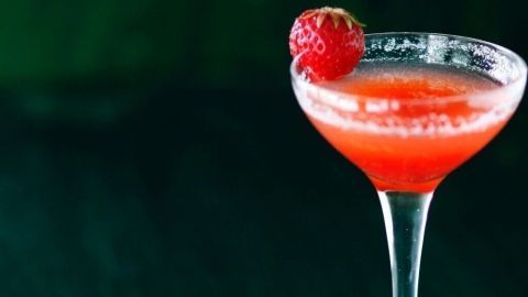 Ein Cocktail in einem eleganten Glas, gefüllt mit rotem Getränk, steht neben frischen Erdbeeren auf einem dunklen Tisch.