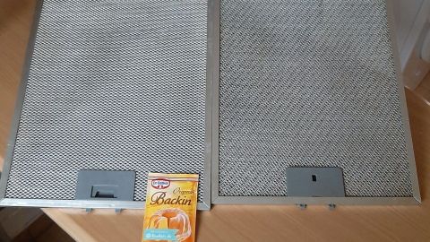 Zwei Dunstabzugshaubenfilter liegen nebeneinander, während ein Päckchen Backpulver am Boden zur Reinigung mit Fettrückständen bereitsteht.