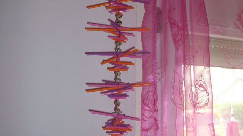 Ein mobiles Kunstwerk aus bunten Trinkhalmen und kleinen Glöckchen hängt in einem Zimmer mit rosafarbenen Vorhängen.