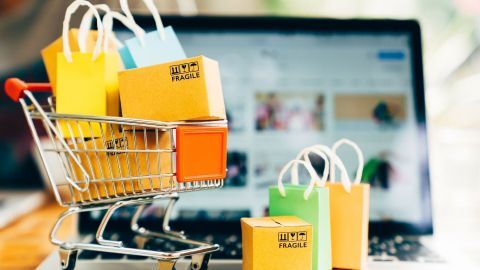 Ein Einkaufswagen steht voll mit bunten Geschenkpäckchen und ist vor einem Laptopbildschirm platziert, der Online-Shopping zeigt.