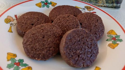 Runde, dunkle Heidesand-Kekse liegen auf einem weihnachtlich dekorierten Teller vor einer Keksdose im Hintergrund.