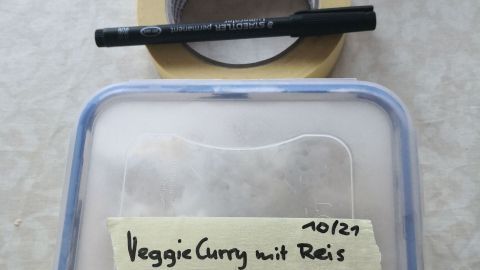 Eine durchsichtige Vorratsdose mit „VeggieCurry mit Reis“ beschriftet, umgeben von Malerkrepp und einem schwarzen Marker.