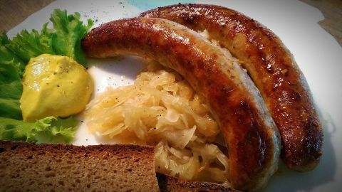 Auf dem Teller liegen zwei goldbraun gebratene Bratwürste, begleitet von Sauerkraut, Senf und einem Salatblatt.