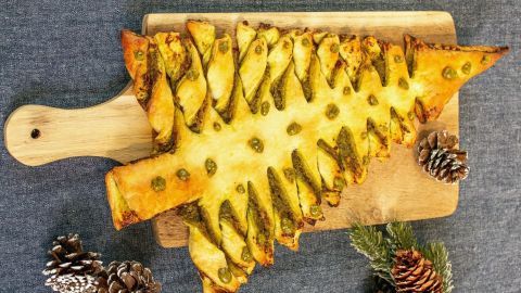 Ein Blätterteig-Tannenbaum mit Pesto liegt auf einem Holzbrett, umgeben von Tannenzapfen und grünen Zweigen.