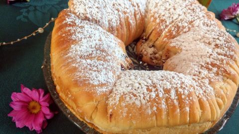 Runder Wollknäul-Brot mit Puderzucker bestäubt, auf einem glänzenden Teller, umgeben von bunten Blumen auf einem Tisch.