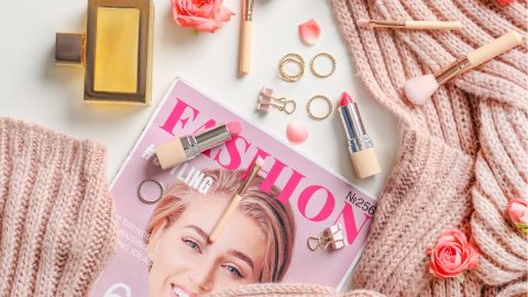 Eine rosa Strickjacke umrahmt eine Modemagazinseite, während Kosmetikprodukte, Ringe und Blumenarrangements darauf liegen.