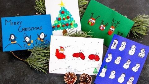 Bunte Weihnachtskarten liegen auf einer dunklen Oberfläche, umgeben von Tannenzweigen und Tannenzapfen, kreativ gestaltet.