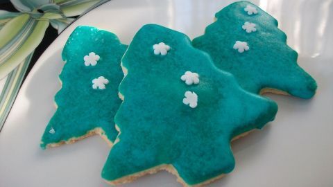 Drei grüne Zuckerplätzchen in Tannenbaumform liegen auf einem weißen Teller und sind mit kleinen weißen Zuckerdeko verziert.