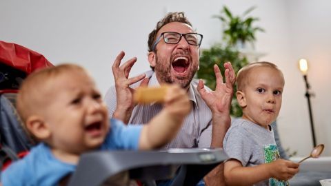 Ein frustrierter Vater schreit laut, während seine beiden weinenden Kinder vor ihm beim Essen sitzen, in einem lebhaften Raum.