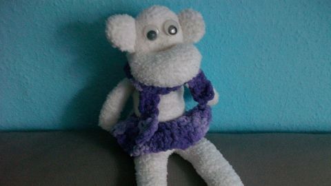 Ein weißes Sockenäffchen sitzt auf einer grauen Oberfläche und trägt ein lila Kleid, vor einer blauen Wand.