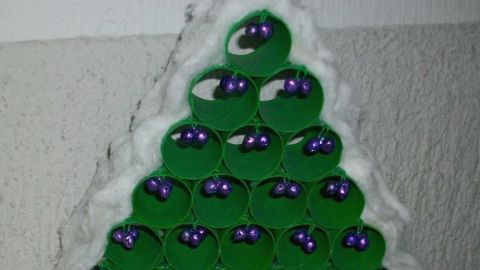 Ein grüner Weihnachtsbaum aus WC-Rollen mit lila Kugeln steht auf einer silbernen Oberfläche, umgeben von einer Wand.