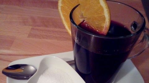 Ein Glas Winzer-Glühwein mit einer Orangenscheibe steht auf einem Teller neben Zucker, umgeben von Holz.