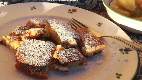 Zarte Bananenschmarren-Stücke, bestäubt mit Puderzucker, auf einem Teller neben einer Schüssel mit fruchtigem Kompott.