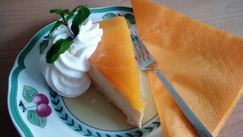 Ein Stück Sanddorn-Torte liegt auf einem dekorativen Teller, neben Schlagsahne und einem orangefarbenen Servietten.