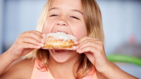 Ein Mädchen beißt genüsslich in einen Zuckerteigkringel, was Fragen über Zucker und Zahngesundheit aufwirft.
